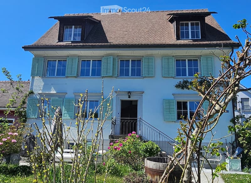 Two-story house with light blue exterior, multiple windows with shutters, and a small garden with flowering plants and a wooden barrel.