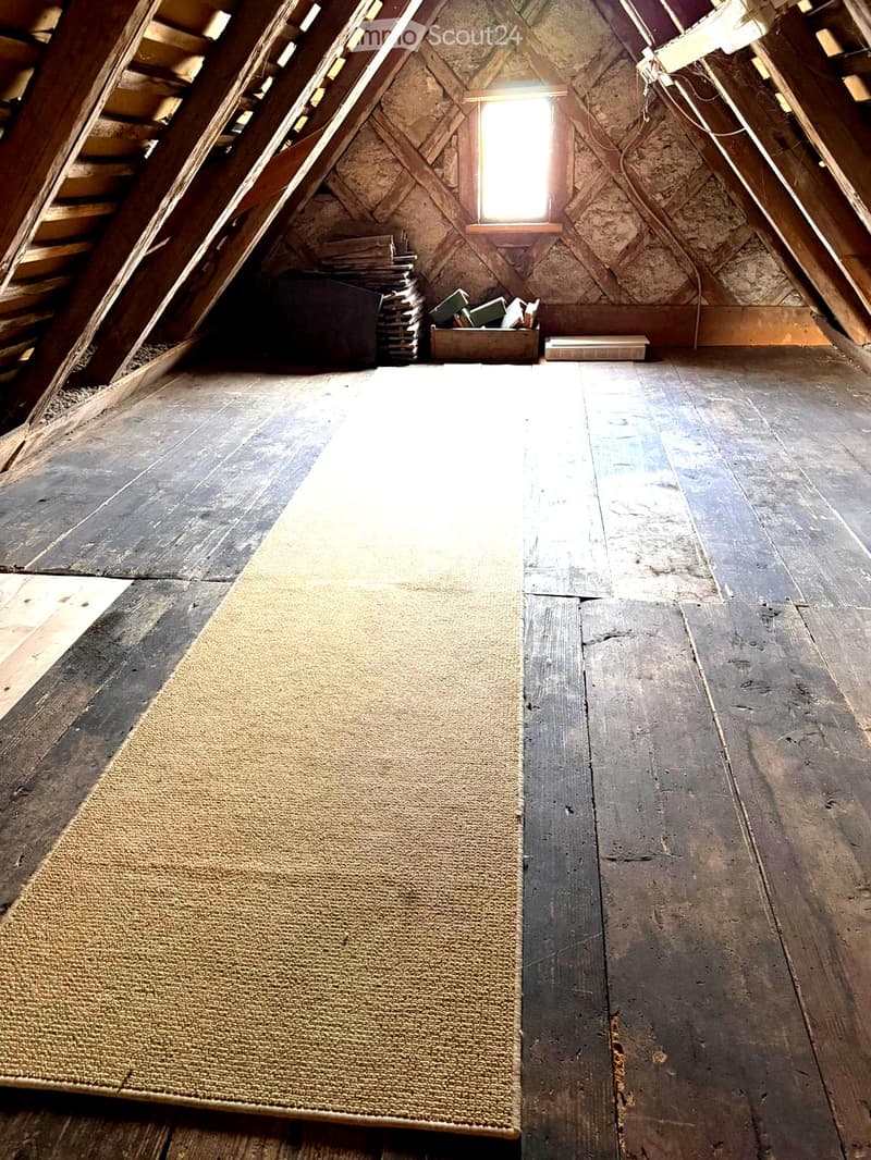 An attic space with exposed wooden beams, slanted wooden walls, and a single window letting in natural light. The floor is made of wide wooden planks, and there is a small pile of items in the corner, including boxes and a small table.