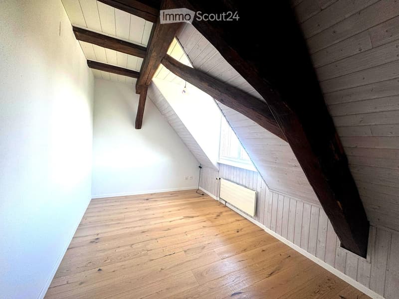 Empty attic room with wooden beams, wooden floor, white walls, and a small window letting in natural light.