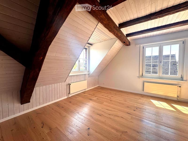 Empty attic room with wooden floors, exposed wooden beams, white walls, and two windows with radiators below them.