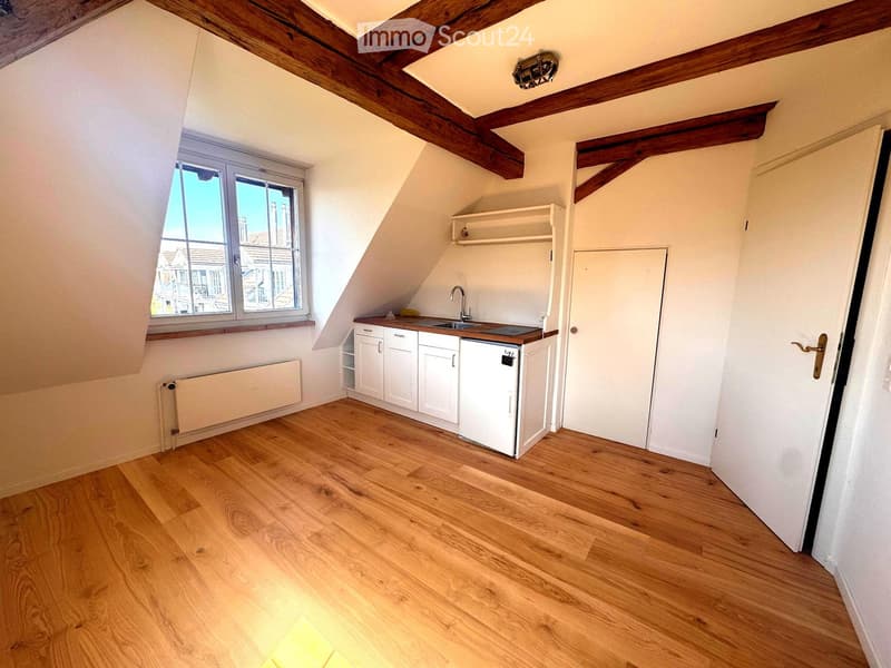 Attic kitchen with wooden floors, white cabinets, a sink, and a refrigerator. Features exposed wooden beams and a large window offering a view of the street.