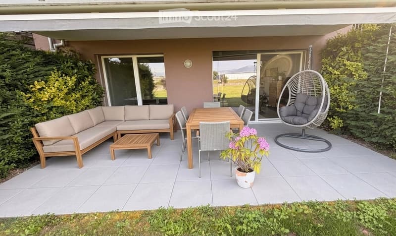 A modern patio area with a grey sectional sofa, wooden coffee table, dining table with chairs, hanging egg chair, and potted plant. Large sliding glass doors lead to the interior, surrounded by greenery and a well-maintained lawn.
