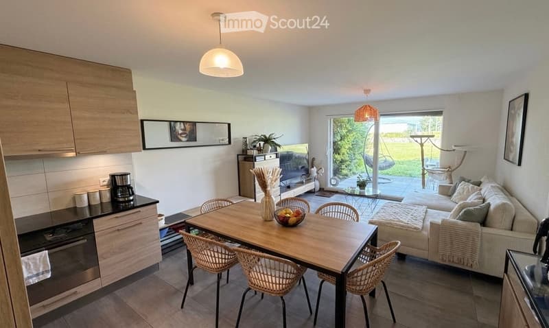 Open-plan living and dining area with modern furniture, a kitchen area, and a view of a garden through large windows.