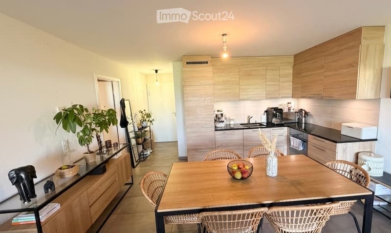 A modern kitchen and dining area with light wooden cabinets, a central dining table with wicker chairs, and various appliances including a coffee maker and oven. There are decorative plants and a vase on the table.