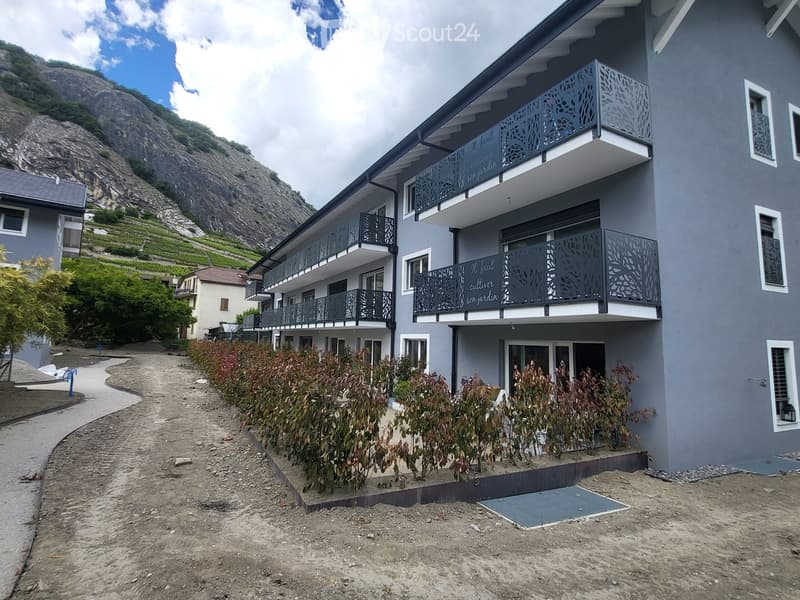 Modern two-story building with grey facade, multiple balconies, and decorative metal railings. Located near a rocky hillside with terraced vineyards.
