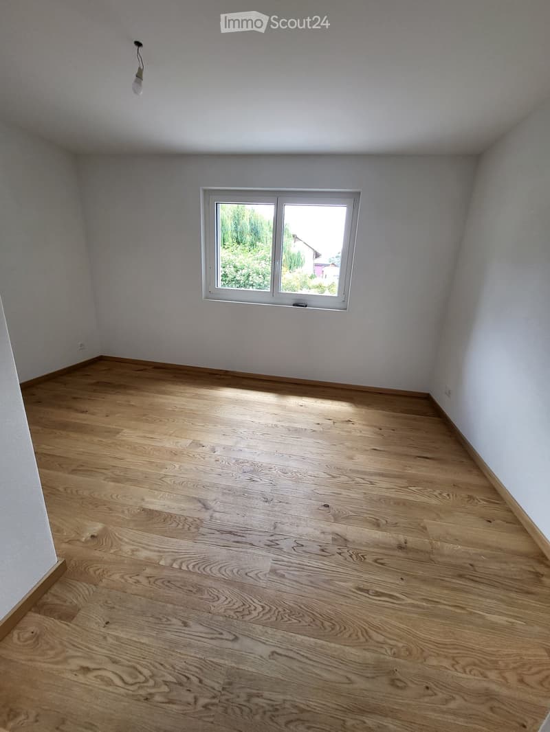 Empty room with light wooden flooring and white walls, featuring a single window with white frame and a ceiling light fixture.