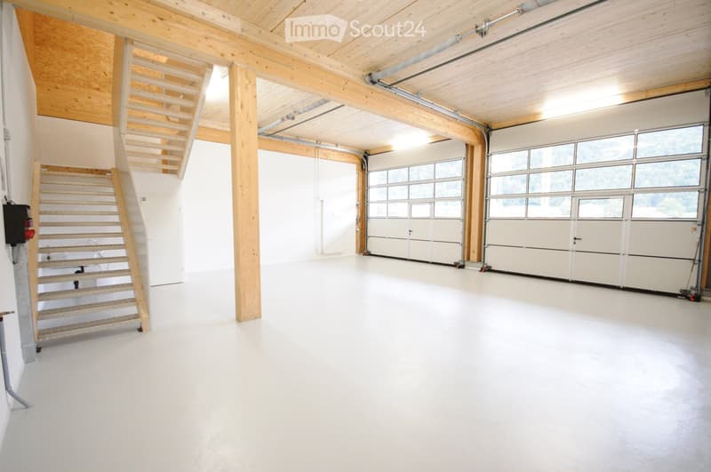 Empty garage with white walls, wooden beams, and large garage doors. A wooden staircase leads to an upper level.