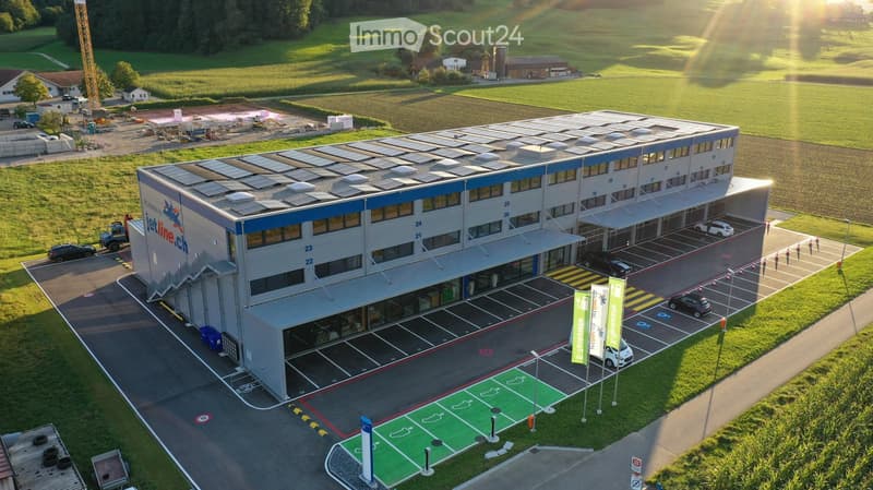 A large building with solar panels on the roof, a parking lot with several cars parked, and a charging station for electric vehicles.