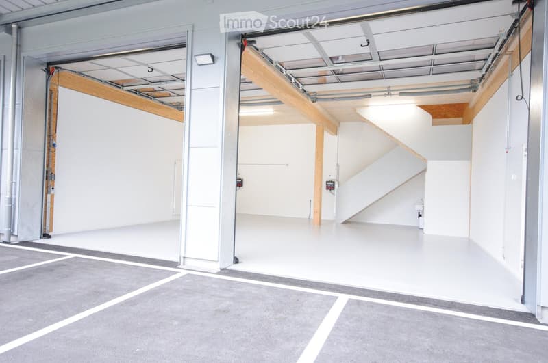Modern industrial-style garage with large open space, white walls, grey flooring, and a staircase leading to an upper level.