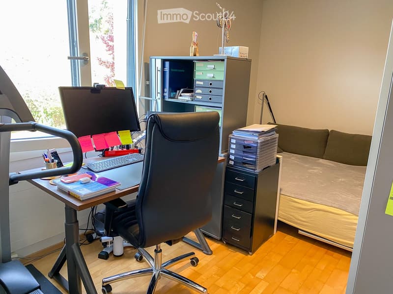 A well-lit room with a desk setup featuring a computer monitor, keyboard, and chair, along with a filing cabinet and a bed in the corner.
