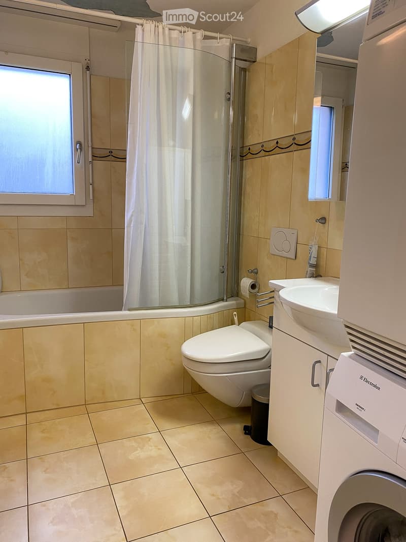 Modern bathroom with beige tiles, bathtub, toilet, sink, shower curtain, and a washing machine.