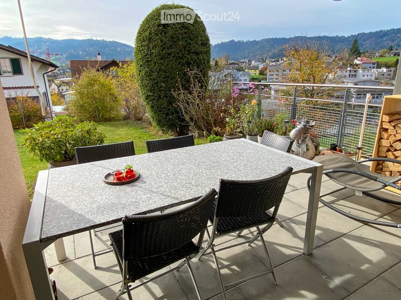Balcony with a round table, four black wicker chairs, and a decorative tray with cups. The balcony overlooks a scenic landscape with trees and houses.