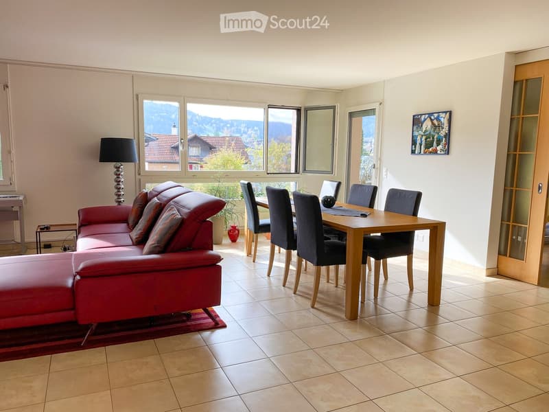 Bright living and dining area with a red leather sofa, wooden dining table, chairs, a floor lamp, and a piano, overlooking a scenic mountain view.