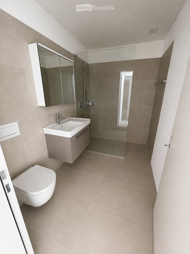 Modern bathroom with beige tiles, white toilet, sink with cabinet, and large mirror above the sink. A glass-enclosed shower area is visible in the background.