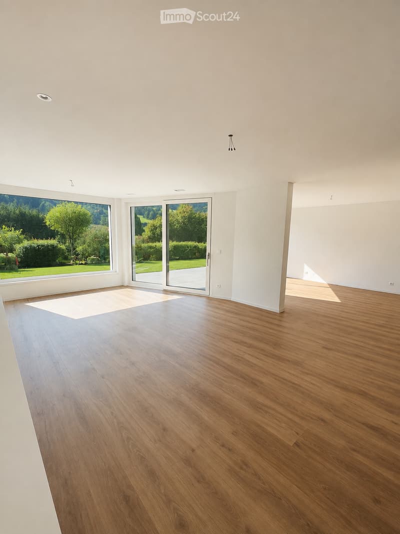 Empty room with wooden flooring, large windows, and sliding glass doors leading to a garden.