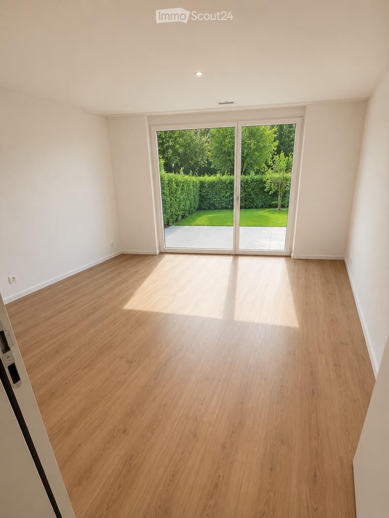 Empty room with light wooden flooring and white walls, featuring a large sliding glass door leading to a green garden.