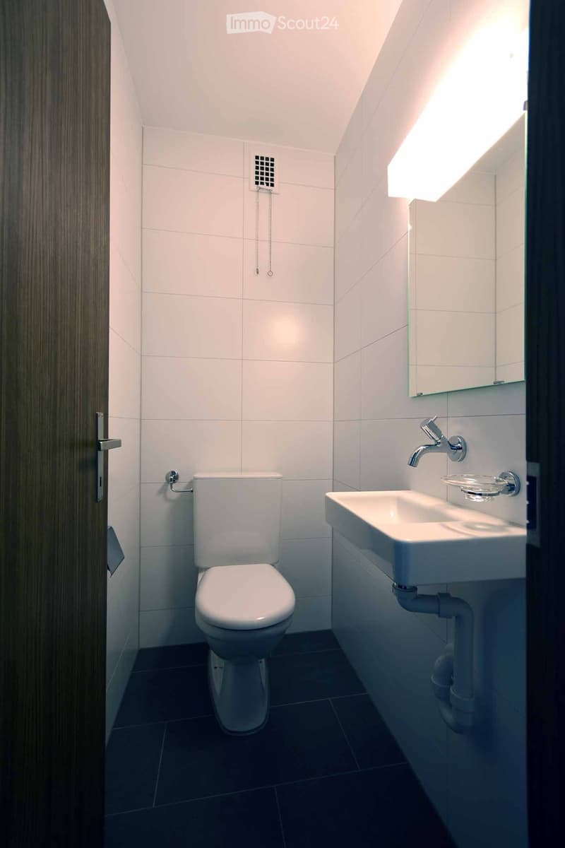 Modern bathroom with white tiles, a toilet, a sink with a modern faucet, and a large mirror with built-in lighting.