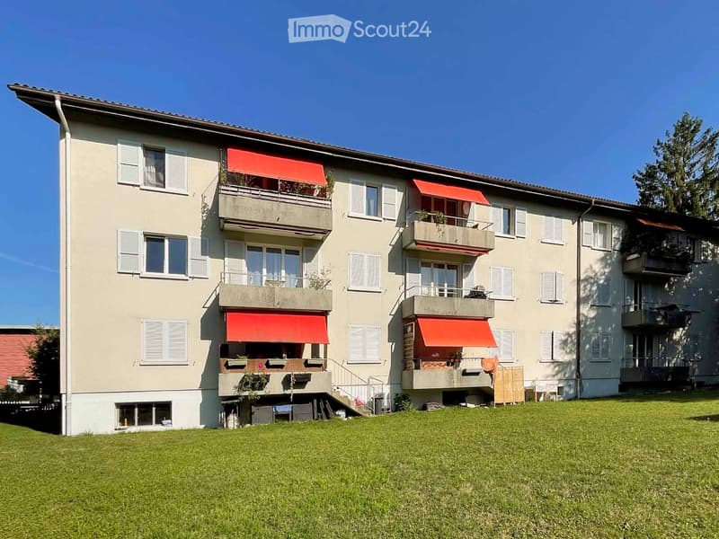 A three-story residential building with balconies, red awnings, and white shutters, surrounded by a grassy lawn.