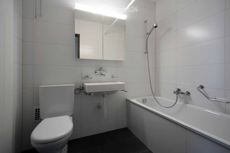 Modern bathroom with white tiles, a toilet, a sink, a bathtub with a handheld shower, and a mirrored cabinet with built-in lighting.