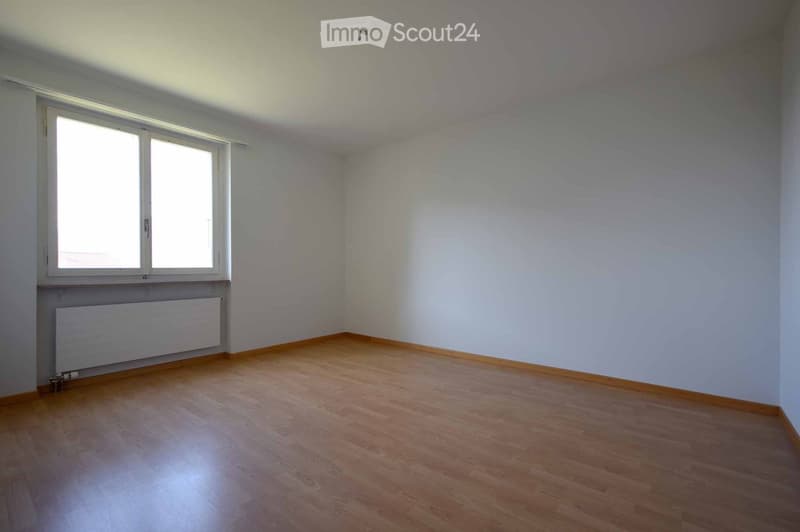 Empty room with wooden flooring, white walls, and a large window with blinds. A radiator is visible under the window.