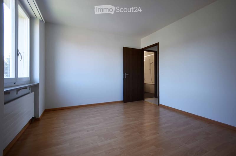 Empty room with wooden flooring, white walls, and a large window. A door leads to a bathroom with a visible bathtub and shower.
