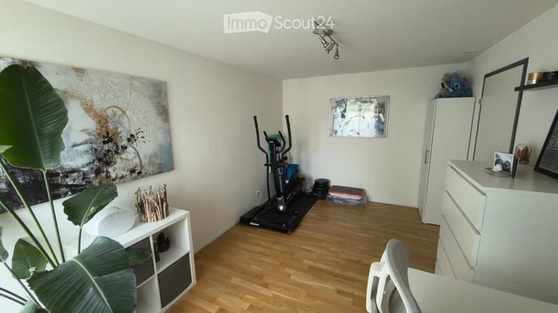 A well-lit room with wooden flooring, a treadmill, a white cabinet, a chair, a shelf with a plant, and artwork on the walls.