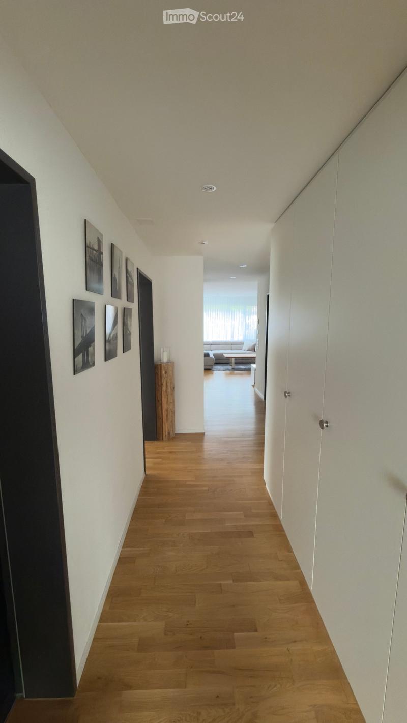 A modern hallway with wooden flooring, white walls, and recessed lighting. Features a small wooden side table with a candle and several framed pictures on the left wall, leading to a living area with a sofa and coffee table visible in the background.