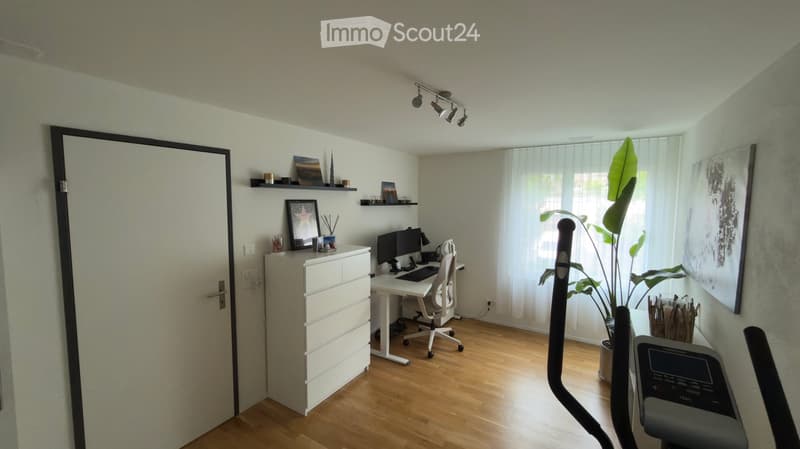 A modern, well-lit room with wooden flooring, a desk setup, a treadmill, and decorative elements including a large plant and artwork.