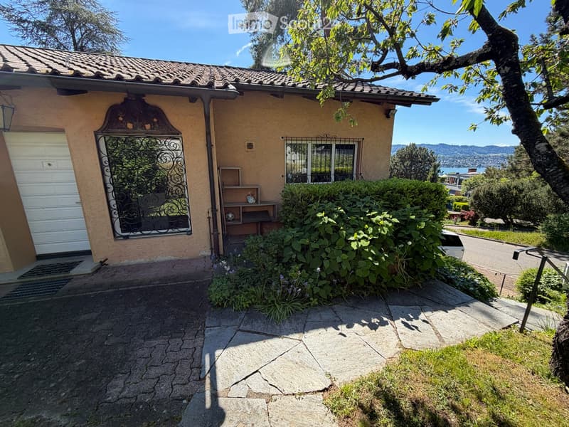 House. Single-story house with beige stucco exterior, tiled roof, white garage door, decorative wrought-iron window, and lush garden with shrubs and flowers.