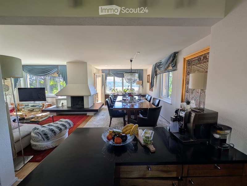 Furnished Room. Open-plan living and dining area with a view, featuring a sofa, coffee table, fireplace, dining table with chairs, television, floor lamp, table lamp, coffee maker, and decorative fruits on a counter.