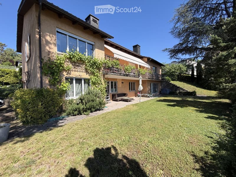 Garden / Outdoor Area. A well-maintained garden with a lawn, surrounded by a two-story house with large windows, a balcony, and climbing plants. Outdoor furniture and a closed umbrella are visible on the patio.