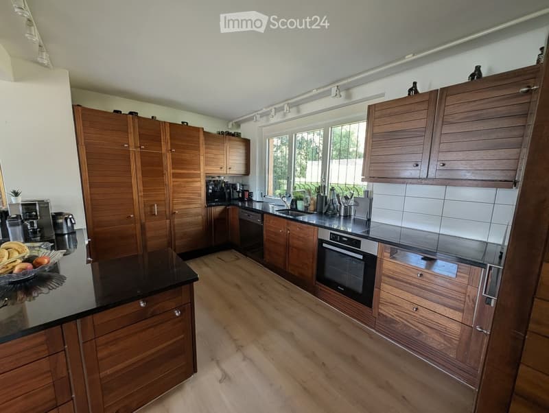 Kitchen. Modern kitchen with wooden cabinets, black countertops, and stainless steel appliances including an oven and dishwasher. The space features a large window with a view of greenery outside.
