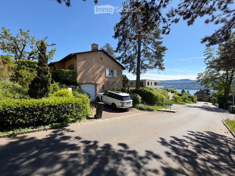 House. A two-story house with a garage, surrounded by greenery and trees, with a driveway leading to the house and a scenic view of a lake and mountains in the background.