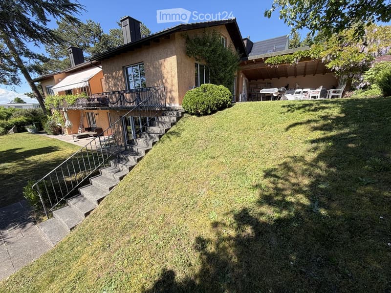 House. Two-story house with a raised ground floor, surrounded by a well-maintained lawn and trees. Features include a staircase leading up to the house, a balcony, and outdoor seating area.