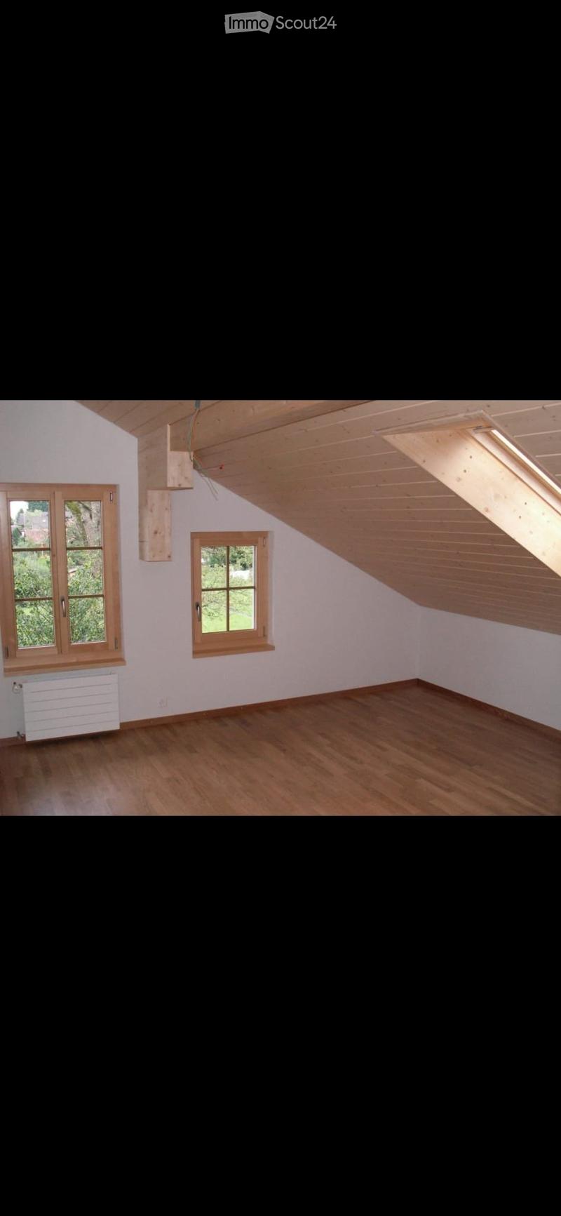Empty attic room with wooden floors, white walls, and two windows with wooden frames. The ceiling has exposed wooden beams and a skylight.