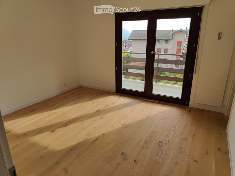 Empty room with light wooden flooring and a large sliding glass door leading to a balcony with a view of the surrounding area.