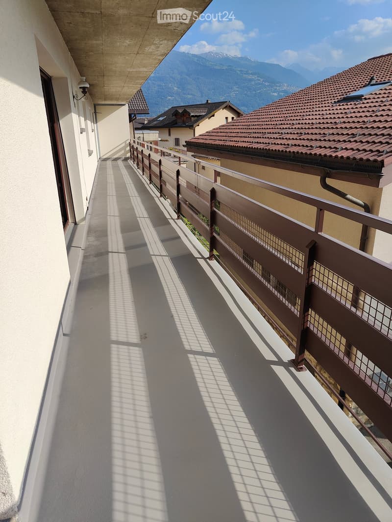 Balcony with metal railing, overlooking a residential area with houses and mountains in the background.