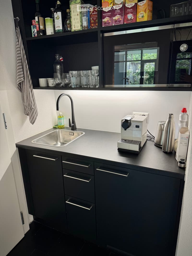 Modern kitchen with dark grey cabinets, stainless steel sink, coffee machine, and various bottles and cups on the counter and shelves.
