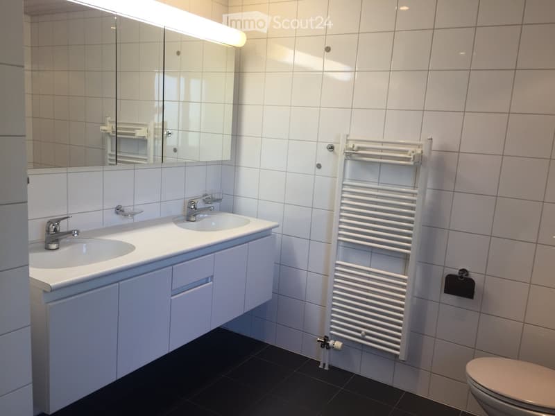 Modern bathroom with white tiles, double sink vanity, mirror, towel rack, and toilet.