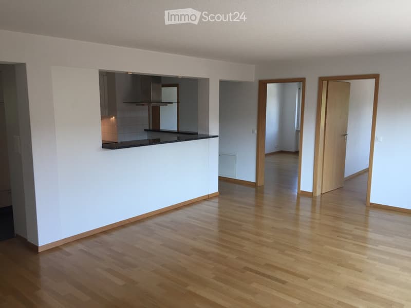Empty room with light wooden flooring, white walls, and a small kitchen area visible in the background. The space is unfurnished with no visible furniture or equipment.