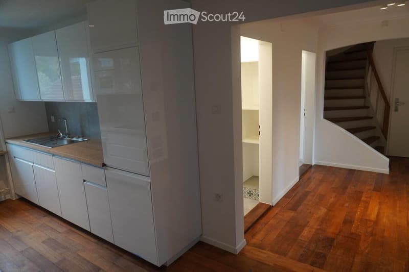 Modern, compact kitchen with white cabinets, a sink, and a wooden floor. An open doorway leads to a staircase area with wooden steps.