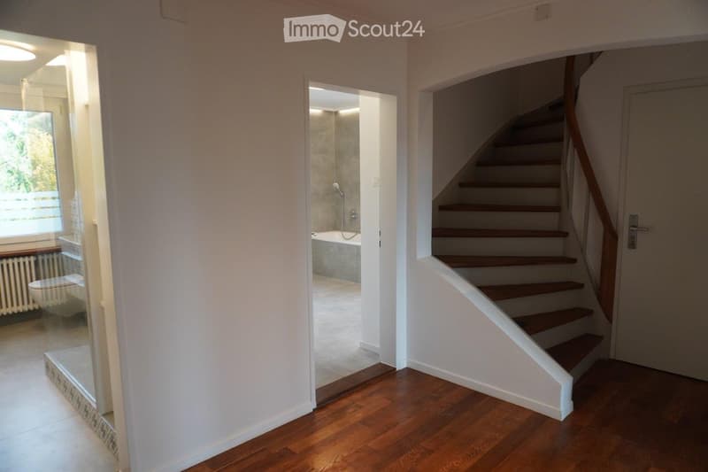 Hallway with wooden flooring, leading to a staircase and a bathroom. The staircase has wooden steps and a wooden handrail. The bathroom is visible through an open doorway, featuring a bathtub, toilet, and a window with blinds.