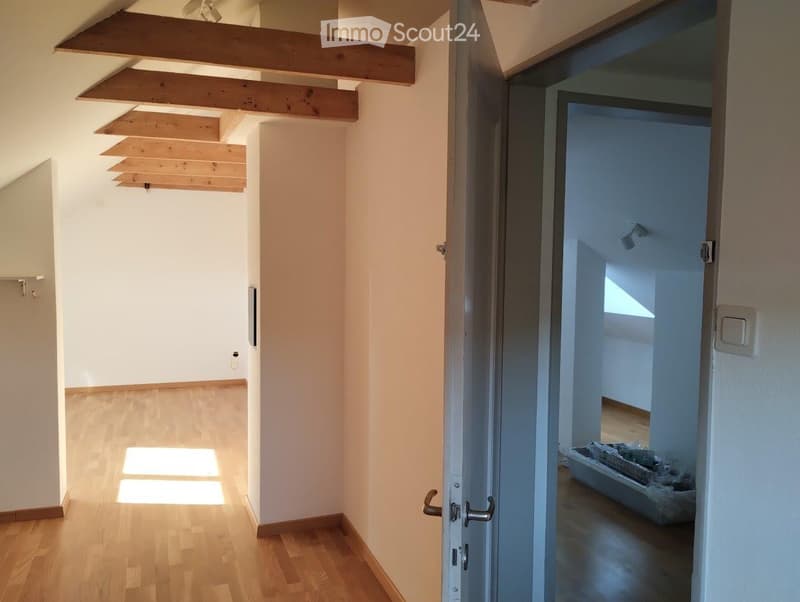 Bright, empty hallway with wooden flooring and white walls, featuring exposed wooden beams on the ceiling and a partially open door leading to another room.