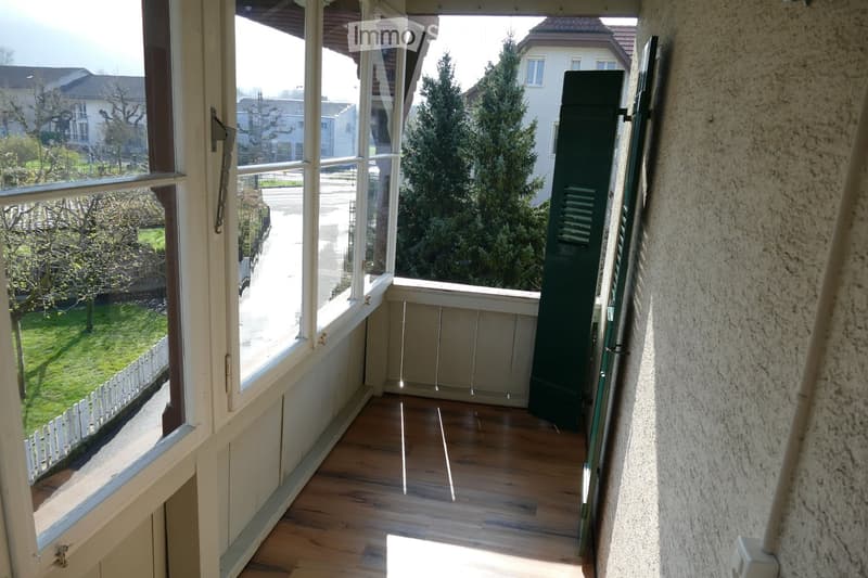 Balcony with wooden flooring, large windows offering views of a residential street and greenery, closed shutters on one side.