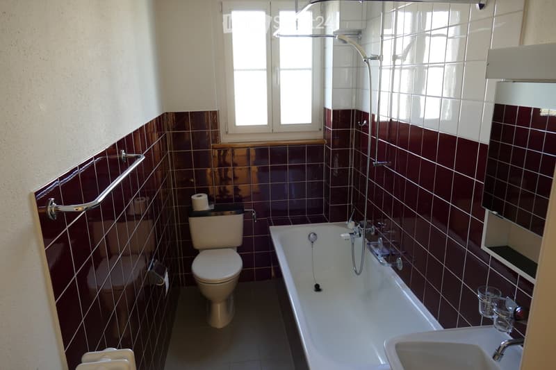 Bathroom with a bathtub, toilet, sink, and mirror. The walls are tiled in dark red and white, and there is a window above the bathtub.