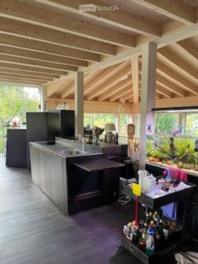 A modern outdoor kitchen area with a wooden deck, exposed beams, and a kitchen island with a countertop, sink, and fridge. There is a small table with various bottles and cups, and a stool near the island.