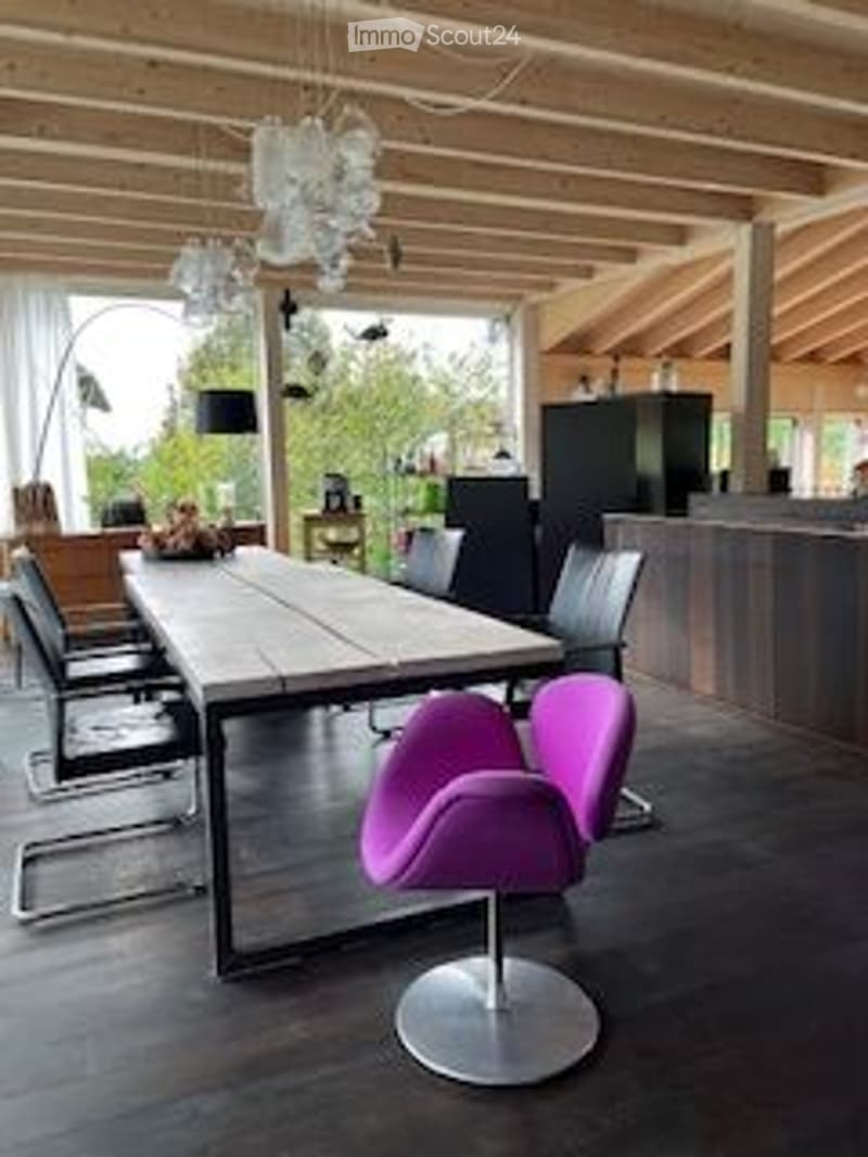 Bright, modern dining area with wooden ceiling beams, a large wooden dining table surrounded by chairs, a purple modern chair, and a sleek kitchen island in the background.