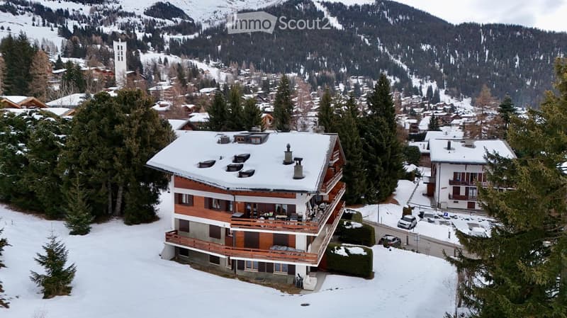 A multi-story wooden chalet with snow-covered roof and balconies, situated in a snowy village surrounded by trees and mountains.