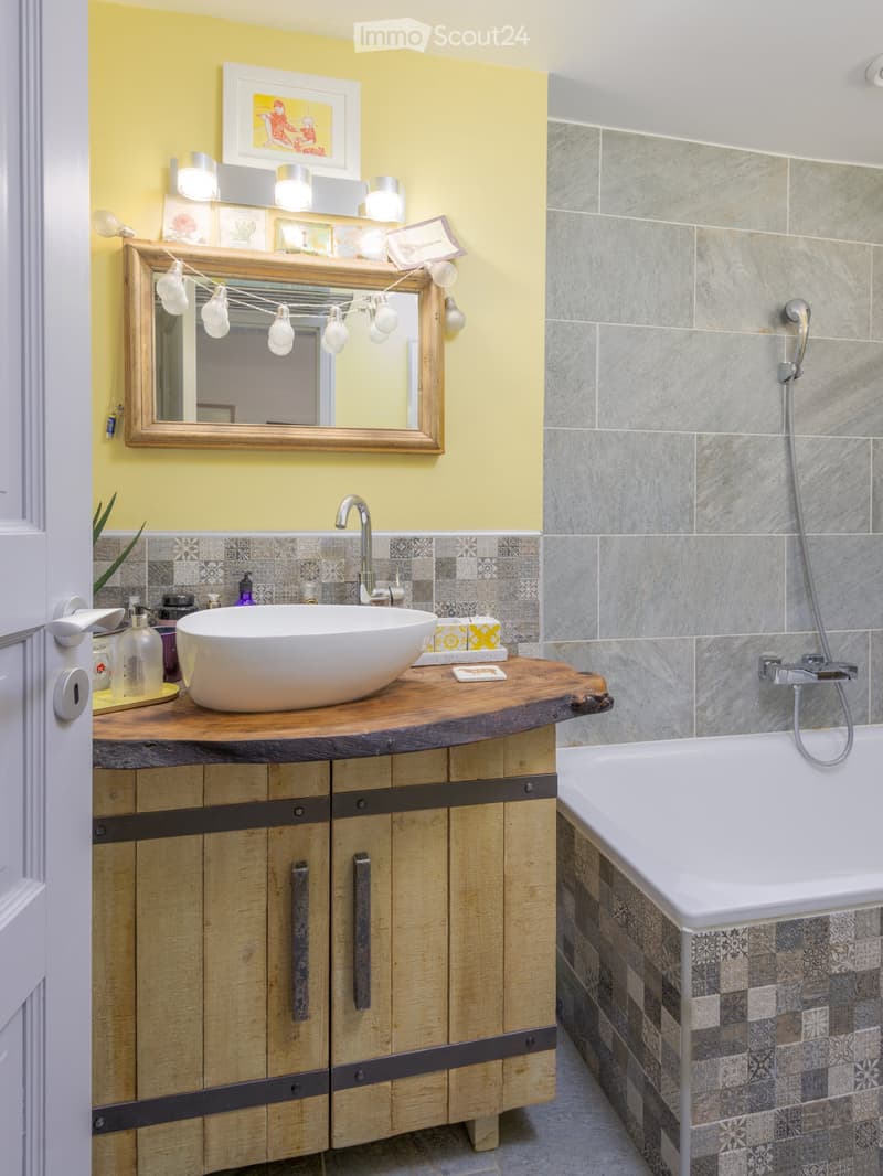 A bathroom with a wooden cabinet under a white sink, a large mirror with a wooden frame, and a bathtub next to it. The walls and floor are tiled with grey tiles, and the bathtub has patterned tiles. There is also a handheld shower and decorative light bul