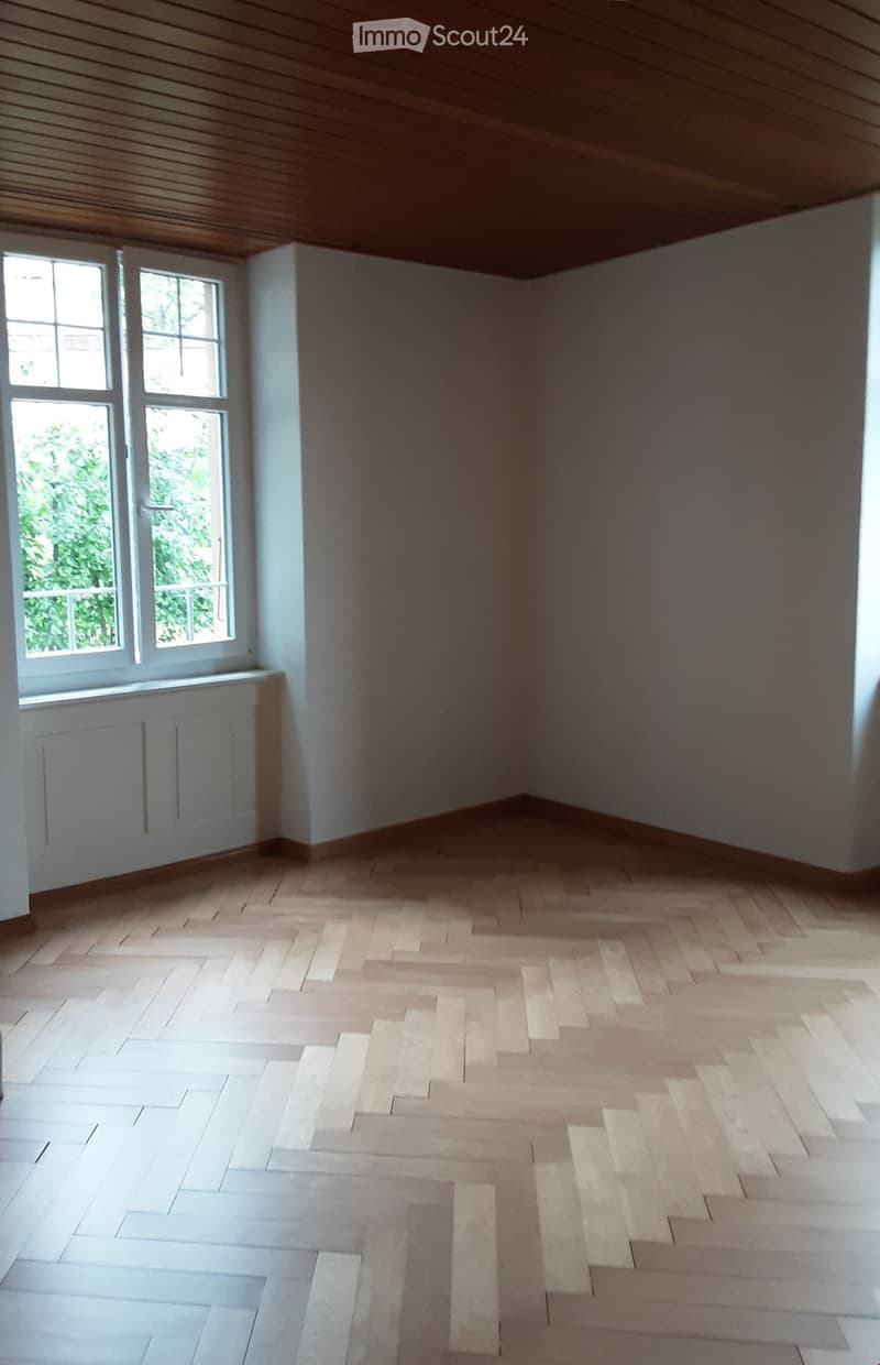 Empty room with light wooden herringbone flooring, white walls, and large windows with white frames. The ceiling is wooden with visible beams.